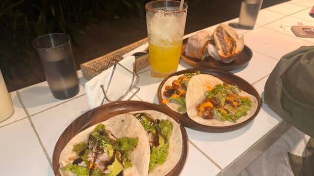 Three plates of tacos and a sandwich with a yellow drink on a white tiled table, ready for a meal in sunny Siargao.
