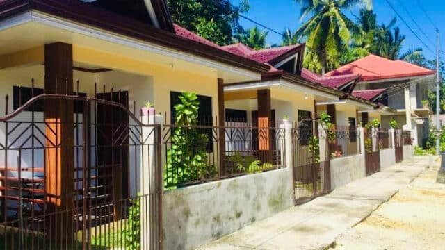 Row of single-story houses with red roofs and gated fences, along a sunny Siargao sidewalk with palm trees nearby.