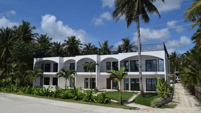 A modern white two-story building with balconies in Siargao, surrounded by palm trees and tropical plants under a blue sky.