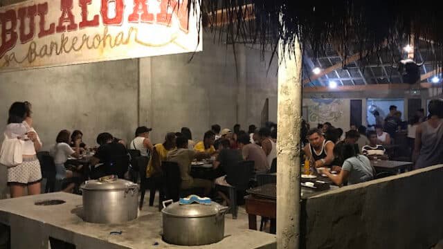 People dining at a casual, open-air Bulaloan in Siargao, with large pots in the foreground and a welcoming island vibe.