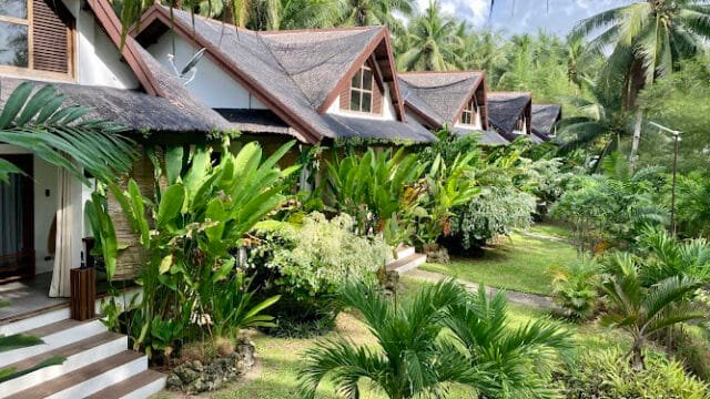 Row of tropical villas in Siargao, surrounded by lush green plants and palm trees under a partly cloudy sky.
