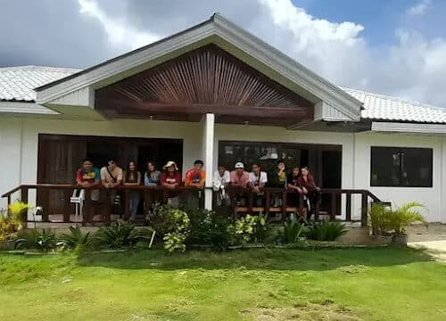 A group of people sit on the porch of a white house in Siargao, with a green lawn and cloudy sky in the background.