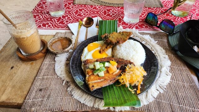 A plate with rice, sunny side eggs, grilled fish, veggies, and sambal—classic Siargao flavors—served with iced coffee and utensils nearby.