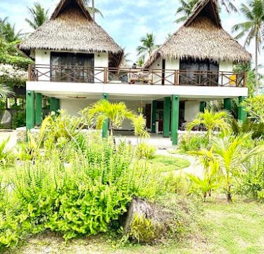 Two-story tropical villa in Siargao with thatched roofs, surrounded by lush green plants and palm trees under a sunny sky.