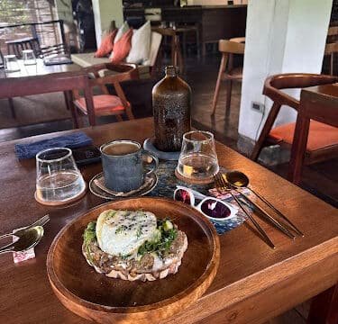 A wooden table with coffee, water, a brown bottle, sunglasses, and food in a cozy Siargao café setting.