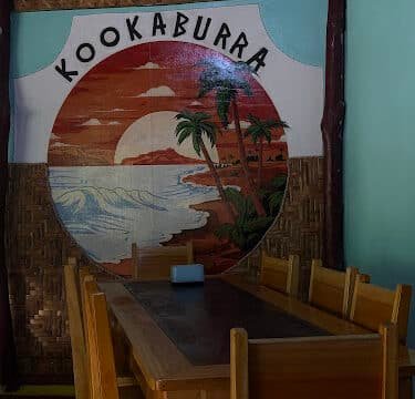 A wooden dining table and chairs sit by a sunset beach mural with Kookaburra and Siargao written above it.
