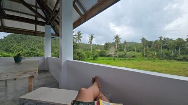 A person relaxes on a terrace with a book, overlooking Siargao’s lush green fields and palm trees under a cloudy sky.
