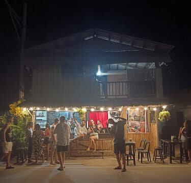 People gather outside a lit wooden house at night in Siargao, some standing and some sitting, enjoying the relaxed atmosphere.