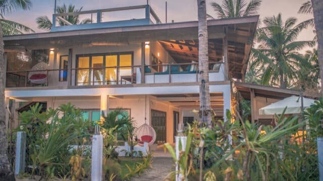 Two-story Siargao beach house with large windows, tropical plants, and palm trees, photographed at sunset.