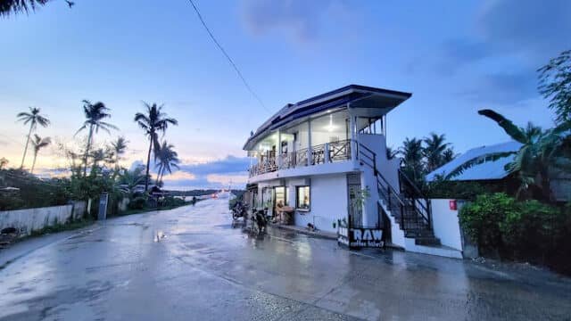 A modern café by a wet road at sunset in Siargao, surrounded by palm trees and a cloudy blue sky.