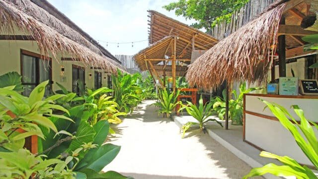 Pathway with sand, tropical plants, thatched-roof huts, and a wooden open-air seating area in bright Siargao sunlight.