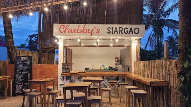 Small open-air food stall Chubbys in Siargao with wooden tables, stools, and string lights at dusk.