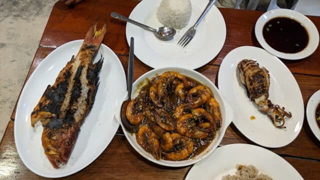 Siargao feast: Grilled fish, sauced shrimp, squid, rice, and dipping sauce on a wooden table; person's hand visible.