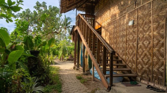 Wooden stairs lead to a bamboo house in Siargao, surrounded by lush green plants; sign says “Remove your footwear.”