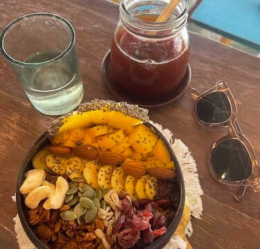 A smoothie bowl with fruit, nuts, and seeds, iced tea in a jar, water glass, and sunglasses on a wooden table in Siargao.