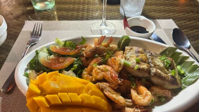 Plate with grilled fish, shrimp, salad, and mango, inspired by Siargao flavors, set on a table with utensils and dipping sauce.