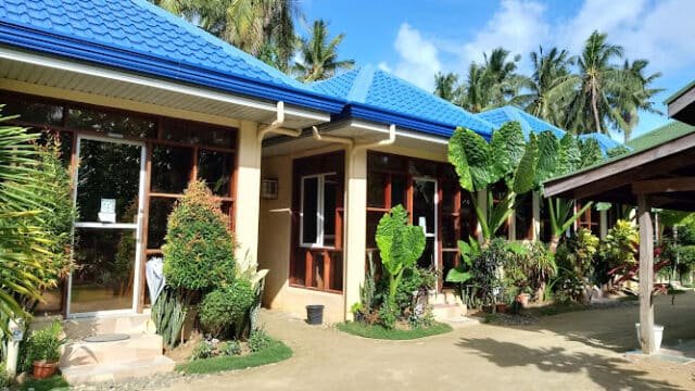 Row of small buildings with blue roofs, large windows, and tropical plants under the sunny Siargao sky.
