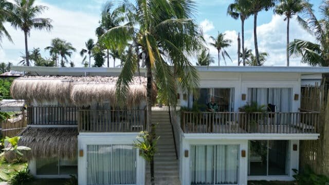 A two-story tropical Siargao villa with palm trees, thatched roof, and balcony, surrounded by lush greenery.