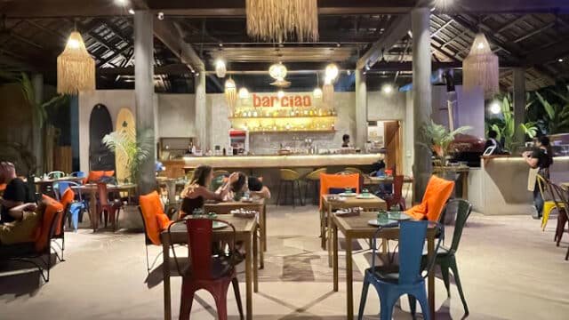 Cozy, rustic Siargao restaurant with colorful chairs, diners at tables, and a warmly lit bar in the background.