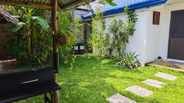 A sunny Siargao backyard with green grass, lush plants, a white house, and a black barbecue grill.