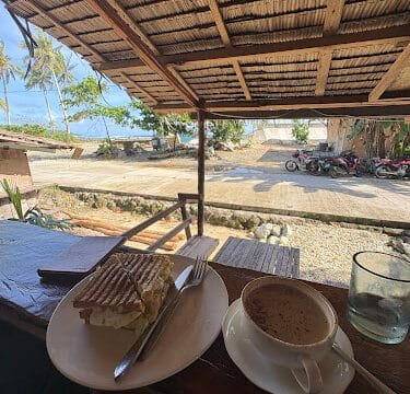A sandwich and coffee on a wooden table with Siargao’s tropical beach and palm trees in the background.