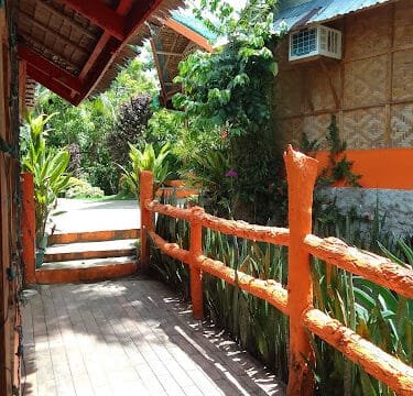 Sunny wooden walkway with an orange railing in Siargao, beside tropical plants and thatched-roof buildings.