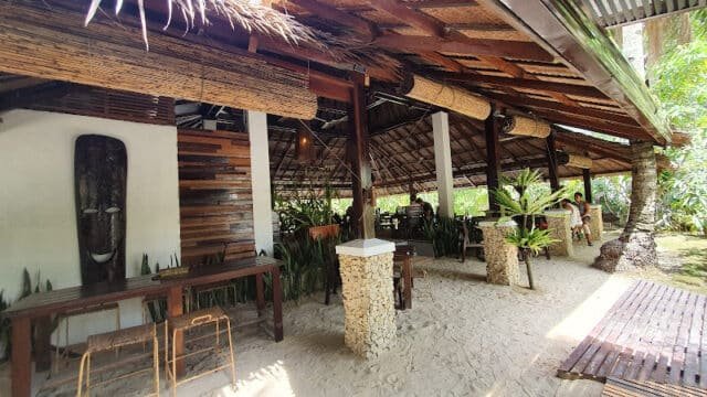 Open-air Siargao beachside restaurant with sandy floor, wooden roof, tropical plants, and rustic decor.