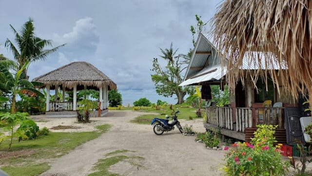A tropical Siargao beach scene with huts, a parked motorcycle, palm trees, and cloudy skies.