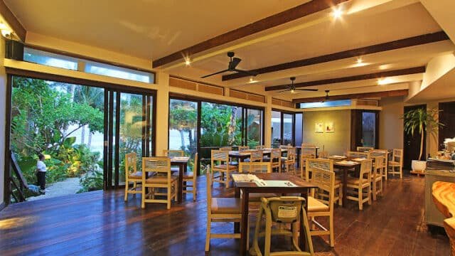 Restaurant with wooden tables and chairs, large windows, and garden views, warmly lit in the evening, capturing the charm of Siargao.
