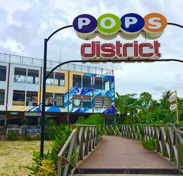 A colorful Pops District sign above a wooden walkway in Siargao, with buildings and lush greenery in the background.