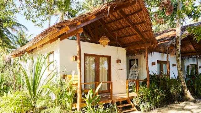 A tropical Siargao bungalow with a thatched roof, hammock, and surrounded by lush greenery and palm trees.