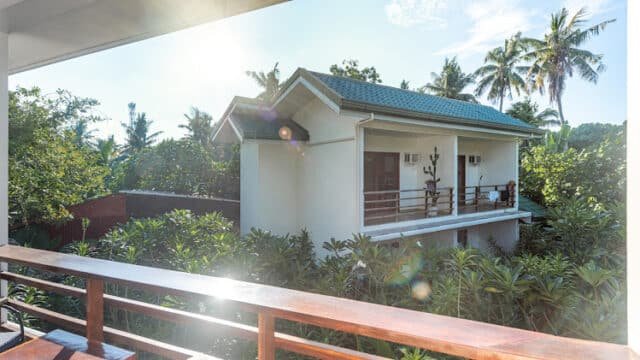Sunlit view of a white house with a balcony in Siargao, surrounded by lush tropical trees and greenery.