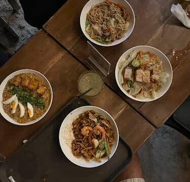 Four bowls of assorted Asian noodle dishes on a wooden table in Siargao, with utensils and a drink nearby.