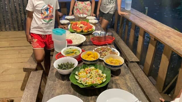Three children stand by a wooden table filled with colorful dishes and drinks, ready for an outdoor meal in vibrant Siargao.
