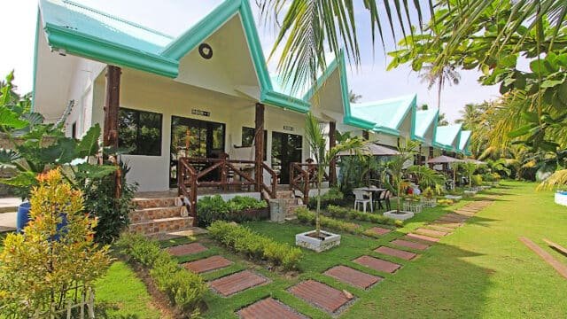 Row of small white bungalows with green roofs, set in a lush Siargao garden and stone path beneath palm trees on a sunny day.