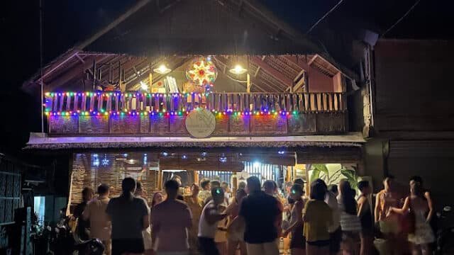 A crowd gathers outside a brightly lit building in Siargao, decorated with colorful string lights at night.