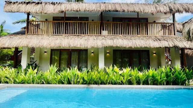 Tropical Siargao villa with thatched roof, balconies, lush plants, and a clear blue swimming pool in front.
