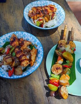 Three plates with grilled chicken skewers, rice, and vegetables on a rustic wooden table in Siargao's natural light.