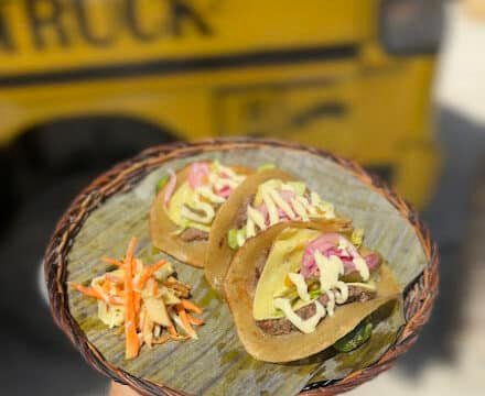 Three tacos with toppings and a side of slaw on a woven platter, held in front of a vibrant Siargao food truck.