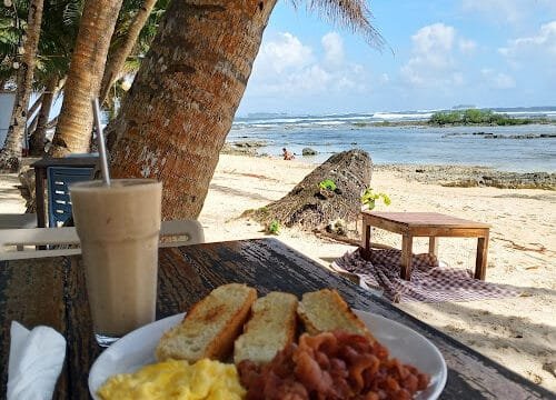 Breakfast plate with toast, scrambled eggs, and bacon on a Siargao beachside table, smoothie in hand, palm trees, and ocean view.