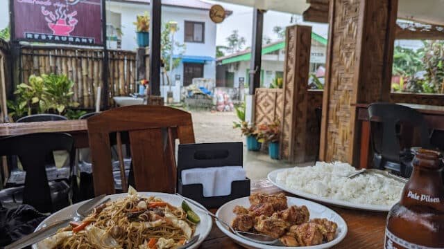 Plates of noodles, fried chicken, rice, and a beer on a wooden table at a Siargao outdoor restaurant.