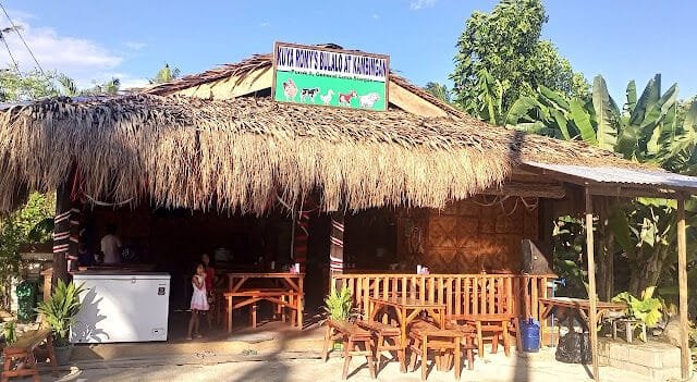 A rustic Siargao hut with a thatched roof, wooden benches, and tropical plants under a bright blue sky.
