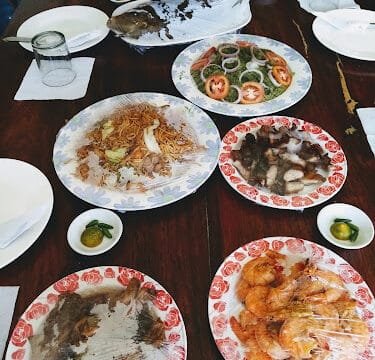 A wooden table set with assorted Filipino dishes, plates, and glasses in Siargao, with food covered in plastic wrap.