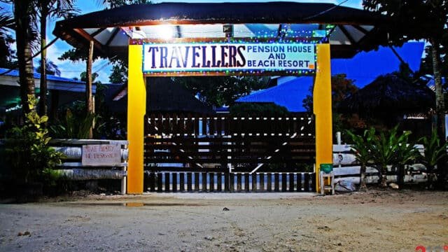 Entrance gate of Travellers Pension House and Beach Resort in Siargao, lit up at dusk with trees and fence in view.
