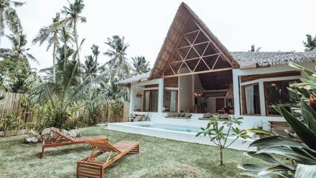 Tropical Siargao villa with a thatched roof, pool, and wooden lounge chairs surrounded by palm trees and lush greenery.