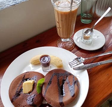 Chocolate pancakes with butter, syrup, banana slices, and iced coffee on a wooden table by a Siargao window.