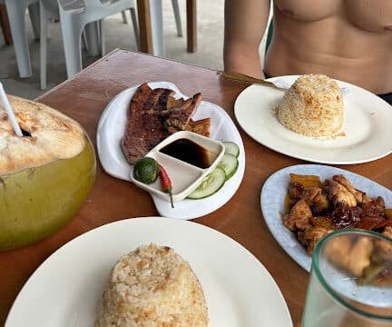A table in Siargao with plated rice, grilled meat, a coconut drink, and a shirtless person sitting at the table.