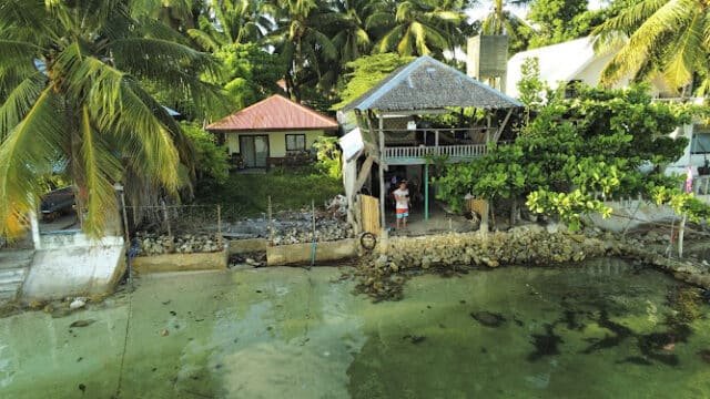 A small seaside house in Siargao with palm trees, a gazebo, and a person standing on a deck by the water.