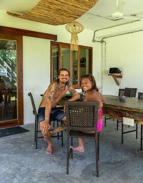 A man and a young girl sit smiling at a table on a covered Siargao patio with wicker chairs and a wooden ceiling decoration.