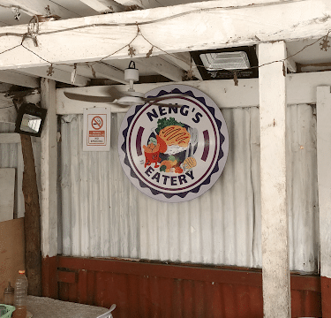 Round sign reading Nengs Eatery with a chicken logo on a rustic wall in Siargao, above plastic chairs and tables.
